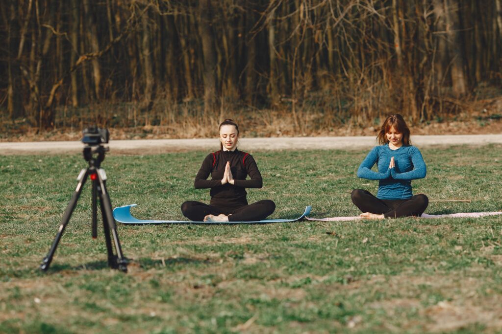 Calm young female friends in sportswear sitting with crossed legs and closed eyes in Namaste pose in front of tripod with photo camera on grass