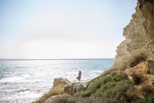 Woman standing in a yoga pose on rocks by the sea, with bright horizon and waves.
